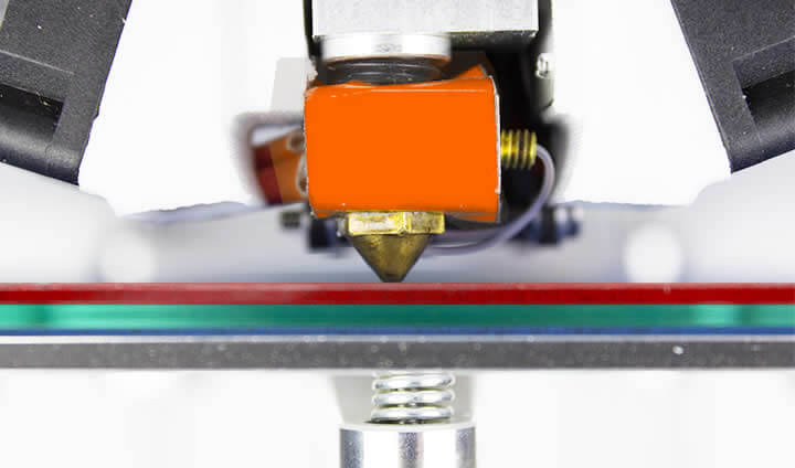 foto com zoom do bico de uma impressora 3d muito próximo da mesa , espremendo o filamento